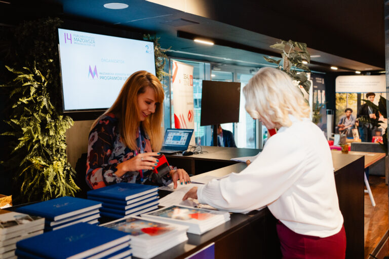 Stanowisko rejestracyjne podczas 14. Forum Rozwoju Mazowsza. Za ladą siedzi kobieta obsługująca uczestników wydarzenia, przed nią stoi inna osoba podpisująca dokumenty. Na biurku leżą stosy materiałów i publikacji, a w tle widać ekran z logotypami organizatora – Mazowieckiej Jednostki Wdrażania Programów Unijnych.