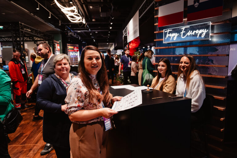 Uczestniczki 14. Forum Rozwoju Mazowsza przy stoisku „Tropy Europy”. Na ladzie widoczna karta z zadaniami i pieczątka, a w tle kolorowe stoiska tematyczne i flagi państw europejskich.