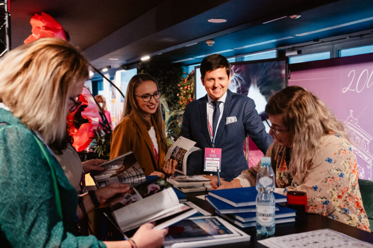 Uczestnicy 14. Forum Rozwoju Mazowsza podczas rozmowy przy stoisku informacyjnym. Na stole widoczne są publikacje i materiały promocyjne wydarzenia, a w tle dekoracje i baner z jubileuszowym motywem „20 lat Mazowsza w UE”