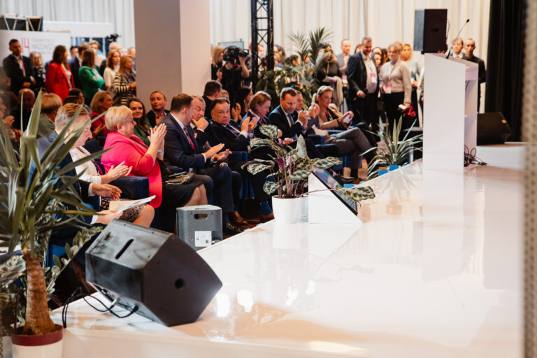 Publiczność 14. Forum Rozwoju Mazowsza podczas jednego z wystąpień na głównej scenie. W pierwszym rzędzie uczestnicy oklaskują prelegenta, a w tle widoczna grupa słuchaczy stojących oraz ekrany i roślinna aranżacja sceny.