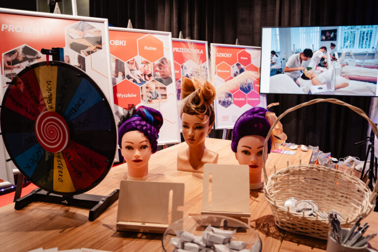 Stoisko Zakładu Doskonalenia Zawodowego w Warszawie podczas 14. Forum Rozwoju Mazowsza. Na stole znajdują się główki fryzjerskie z fantazyjnymi fryzurami oraz koło fortuny z zadaniami i nagrodami, a w tle widoczne są plakaty prezentujące działalność edukacyjną ZDZ.