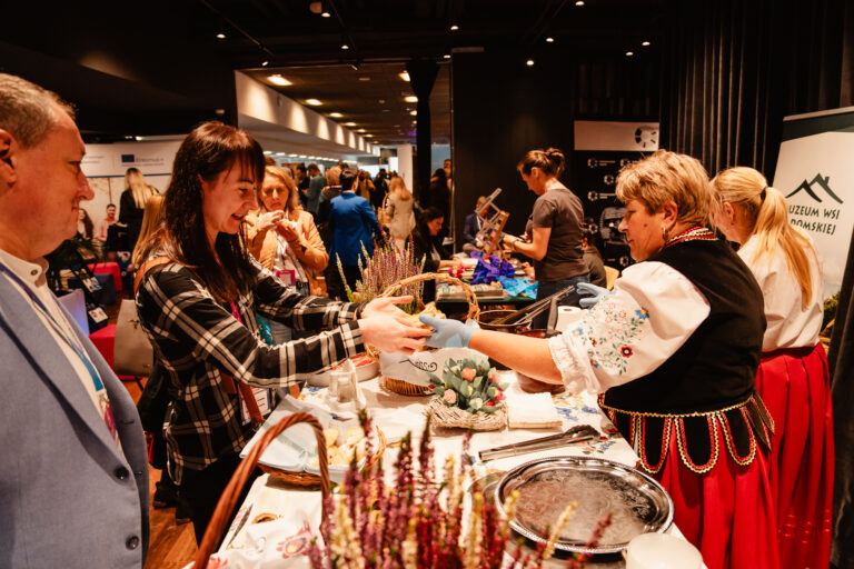 ChatGPT powiedział: Stoisko Muzeum Wsi Radomskiej podczas 14. Forum Rozwoju Mazowsza. Kobiety w tradycyjnych strojach ludowych prezentują regionalne potrawy i wyroby, częstując uczestników wydarzenia.