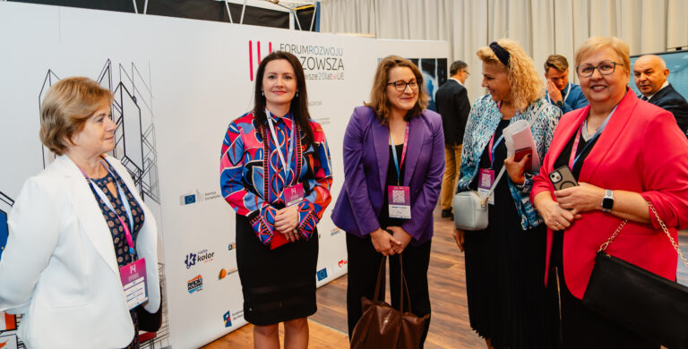 Grupa kobiet uczestniczących w 14. Forum Rozwoju Mazowsza stoi przy ściance z logotypami wydarzenia i partnerów. Uczestniczki rozmawiają i uśmiechają się, trzymając identyfikatory oraz materiały konferencyjne. W tle widoczni są inni goście wydarzenia.