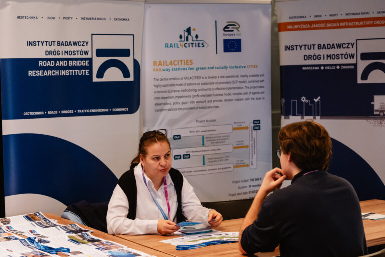 Stoisko Instytutu Badawczego Dróg i Mostów podczas 14. Forum Rozwoju Mazowsza. Przedstawicielka instytutu rozmawia z uczestnikiem wydarzenia o projektach badawczych, w tym o inicjatywie RAIL4CITIES, poświęconej zrównoważonemu rozwojowi stacji kolejowych w Europie