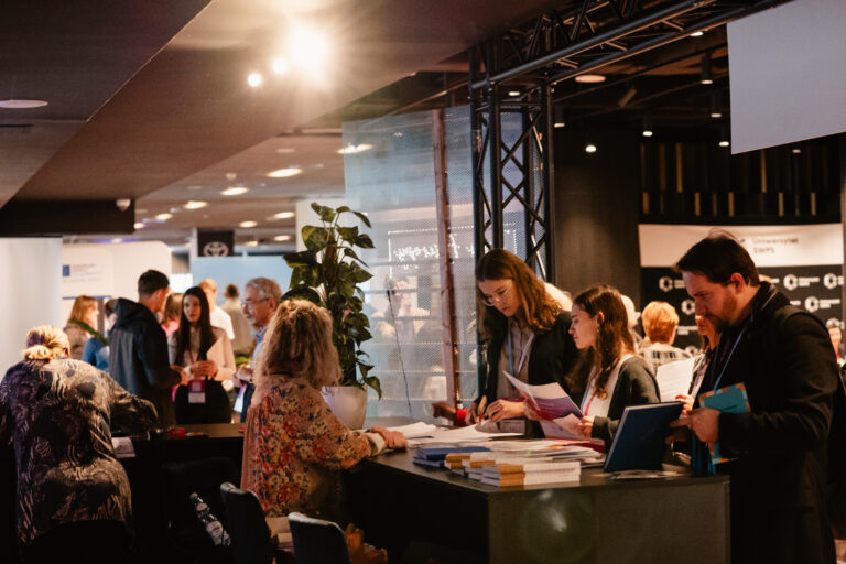 Uczestnicy 14. Forum Rozwoju Mazowsza odwiedzają stoiska wystawiennicze, przeglądając publikacje i materiały informacyjne. W tle widać rozmowy i wymianę doświadczeń między przedstawicielami instytucji i gośćmi wydarzenia.