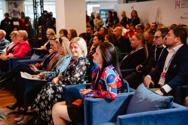 „Na zdjęciu widać uczestników Forum Rozwoju Mazowsza siedzących w niebieskich fotelach i słuchających wystąpienia. W pierwszym rzędzie znajduje się kilka osób ubrane elegancko, z identyfikatorami wydarzenia na szyjach. W tle widoczni są kolejni uczestnicy oraz baner z logotypem Forum.