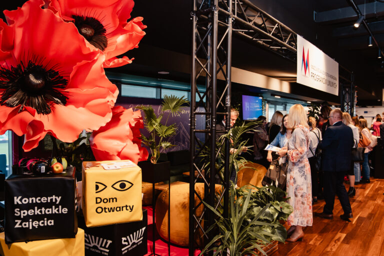 Zdjęcie przedstawia stoisko Mazowieckiej Jednostki Wdrażania Programów Unijnych na Forum Rozwoju Mazowsza. Wystawa ozdobiona jest dużymi, dekoracyjnymi czerwonymi kwiatami i zielonymi roślinami. Na pierwszym planie widać kostki z napisami ‘Koncerty, Spektakle, Zajęcia’ oraz ‘Dom Otwarty’. W tle uczestnicy wydarzenia rozmawiają i oglądają materiały informacyjne.