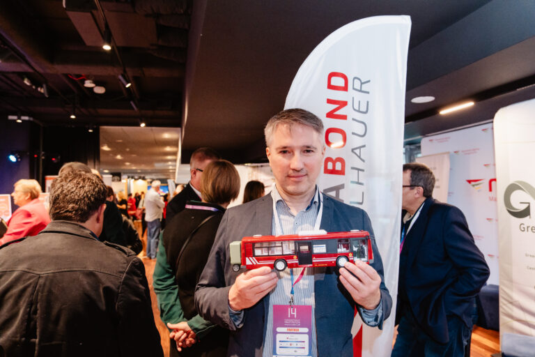 Na zdjęciu mężczyzna stoi na tle stoiska z banerem i trzyma w dłoniach czerwony model autobusu. Wokół niego znajdują się uczestnicy wydarzenia Forum Rozwoju Mazowsza, rozmawiający i oglądający prezentacje wystawców