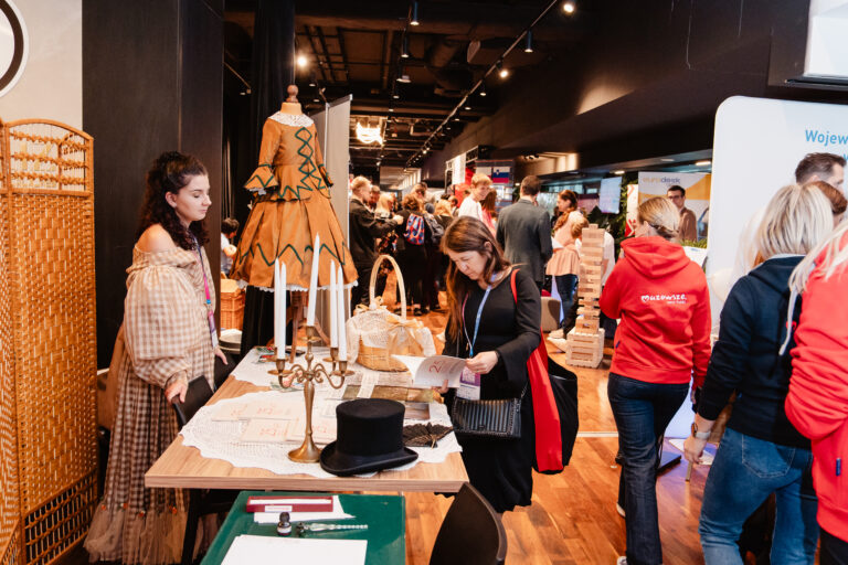 Na zdjęciu stoisko Muzeum Romantyzmu w Opinogórze podczas wydarzenia Forum Rozwoju Mazowsza. Po lewej stronie kobieta w stylizowanym stroju z epoki stoi obok manekina z suknią w kolorze miodowym ozdobioną zielonymi wstawkami. Na stole znajdują się świece, cylinder i broszury. Obok uczestniczka wydarzenia przegląda katalog. W tle widać innych odwiedzających i stoiska partnerów wydarzenia.