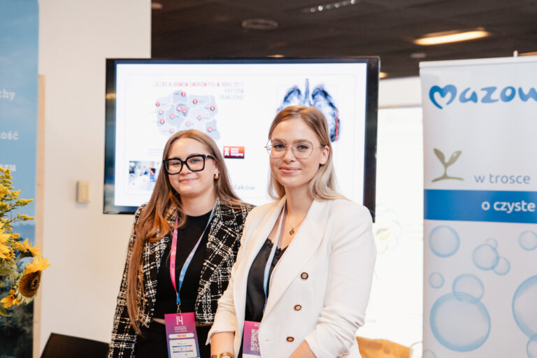 Dwie kobiety stoją przy stoisku informacyjnym podczas wydarzenia Forum Rozwoju Mazowsza. W tle widoczne są banery i ekran z prezentacją dotyczącą projektów zdrowotnych i ekologicznych Mazowsza