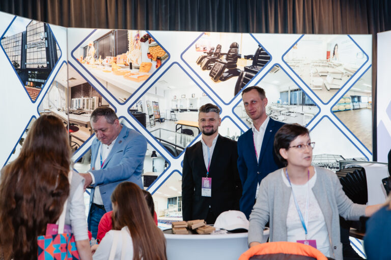 Alt: Stoisko Wyższej Szkoły Inżynierii Zdrowia, przy którym kilka osób rozmawia i prezentuje materiały promocyjne na tle plansz z fotografiami sal dydaktycznych i wyposażenia uczelni.