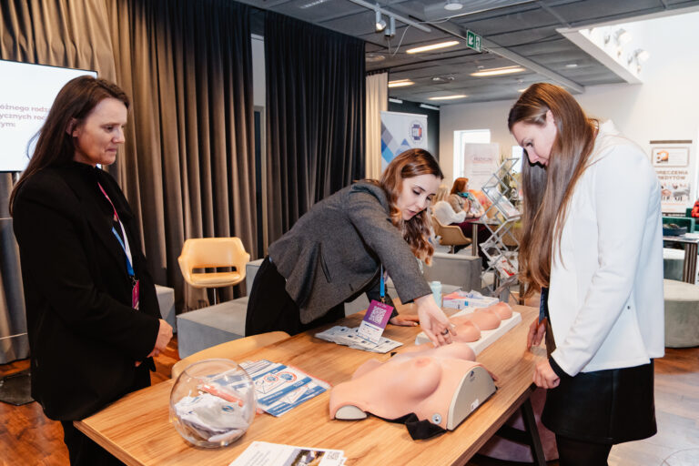 Trzy kobiety stoją przy stole z modelami piersi, ucząc się samobadania w ramach warsztatów edukacyjnych dotyczących profilaktyki zdrowia.