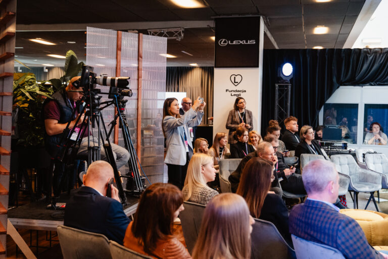 Na zdjęciu widać część publiczności siedzącej w sali konferencyjnej podczas Forum Rozwoju Mazowsza. Po lewej stronie stoi operator kamery nagrywający wydarzenie, obok niego osoba robi zdjęcia telefonem. W tle widać banery z logotypami Fundacji Legii i marki Lexus oraz grupę uczestników słuchających wystąpienia.