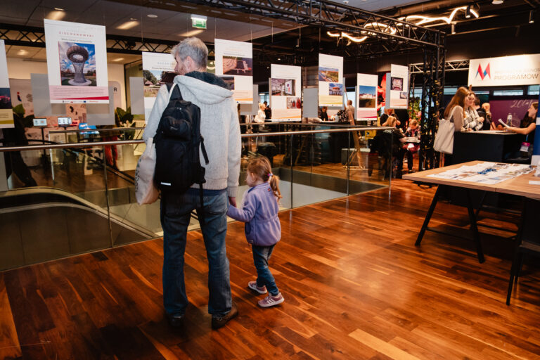 Meżczyzna z plecakiem i mała dziewczynka oglądają wystawę plansz z fotografiami i opisami projektów w nowoczesnym wnętrzu centrum wydarzeń