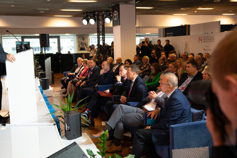 Publiczność podczas inauguracji 14. Forum Rozwoju Mazowsza, siedząca przed główną sceną i słuchająca wystąpienia otwierającego. Na zdjęciu widać uczestników wydarzenia, przedstawicieli instytucji i organizatorów, a w tle fotoreporterów i obsługę techniczną.