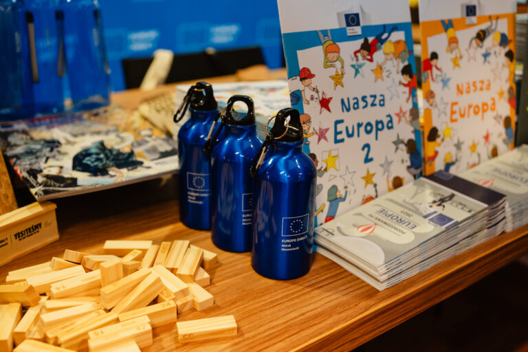 Stoisko Europe Direct Mińsk Mazowiecki. Na stole ustawiono niebieskie butelki z logo Unii Europejskiej, publikacje informacyjne „Nasza Europa” oraz materiały edukacyjne o Unii Europejskiej. Obok znajdują się drewniane klocki edukacyjne.