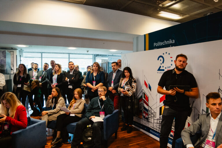 Uczestnicy 14. Forum Rozwoju Mazowsza stojący i siedzący w sali konferencyjnej podczas jednej z prezentacji. W tle widoczna ścianka z grafiką promującą 20 lat Polski w Unii Europejskiej oraz logotypem Politechniki Warszawskiej.
