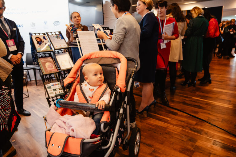 Uczestnicy 14. Forum Rozwoju Mazowsza odwiedzają jedno ze stoisk wystawowych. Na pierwszym planie widoczne dziecko w wózku, a w tle osoby rozmawiające przy ekspozycji z materiałami informacyjnymi i publikacjami.