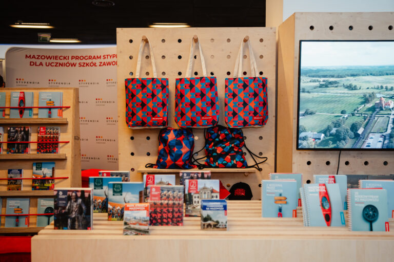 Stoisko promujące Mazowiecki Program Stypendialny dla uczniów szkół zawodowych. Na półkach i stojakach znajdują się publikacje i materiały informacyjne o Mazowszu, a na ścianie wiszą torby i worki w czerwono-niebieski wzór z logo Mazowsze. Obok ekran prezentuje nagrania z regionu.