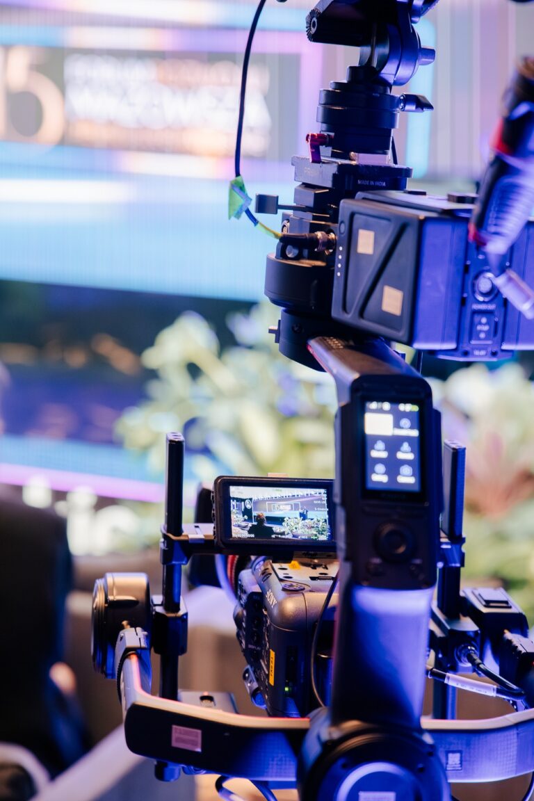 Profesjonalna kamera telewizyjna ustawiona na statywie filmuje wydarzenie, w tle widoczny rozmyty ekran sceniczny i rośliny dekoracyjne.