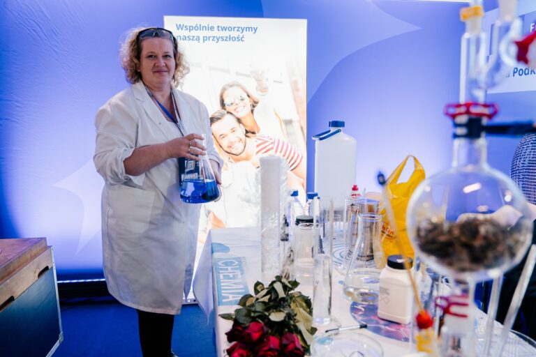 Stoisko województwa podlaskiego; kobieta w białym fartuchu laboratoryjnym prezentuje kolbę z niebieską cieczą, na stole obok znajduje się sprzęt chemiczny i odczynniki.