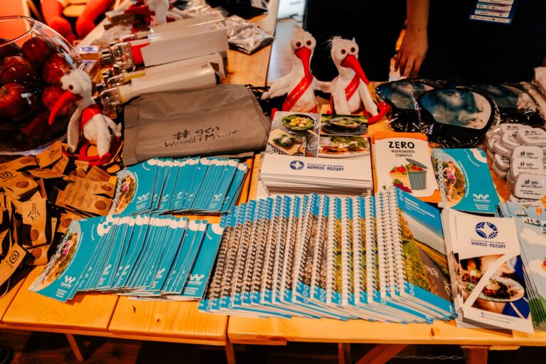 Stoisko promocyjne Warmii i Mazur; na stole ułożone broszury, notesy, ulotki i gadżety z logo regionu, w tym maskotki bocianów i torba z napisem „#gen Warmii i Mazur”.