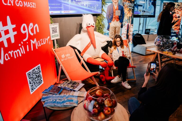 Stoisko Warmii i Mazur; kobieta siedzi na leżaku obok dużej maskotki bociana, uśmiecha się do zdjęcia robionego telefonem. Wokół widoczne materiały promocyjne i jabłka w szklanej misce.