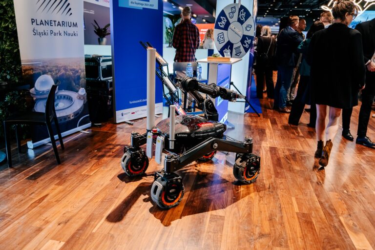 Stoisko Planetarium – Śląskiego Parku Nauki; na pierwszym planie stoi czarny robot mobilny z ramieniem, w tle widoczne banery informacyjne i odwiedzający wydarzenie.