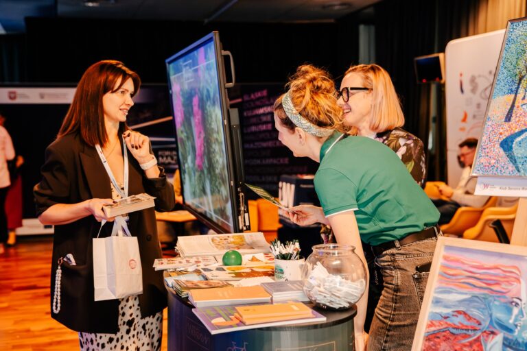 Stoisko Dzielnicy Wawer; trzy kobiety rozmawiają przy ekranie dotykowym, na stole leżą broszury i gadżety promocyjne.