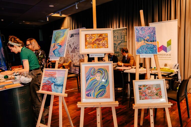 Stoisko Dzielnicy Wawer z kolorowymi obrazami na sztalugach; w tle osoby wykonujące prace plastyczne przy stolikach z materiałami.
