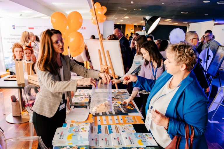 Na zdjęciu widać stoisko z materiałami promocyjnymi i broszurami. Po lewej stronie kobieta w okularach przekazuje coś do torby kobiecie po prawej. W tle stoją inne osoby oraz pomarańczowe balony. Panuje atmosfera targów lub wydarzenia wystawienniczego.