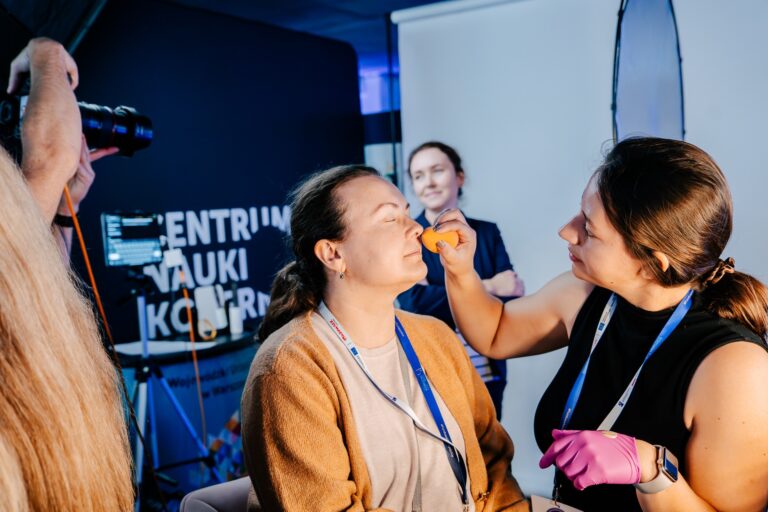 Na zdjęciu widać przygotowania do profesjonalnej sesji zdjęciowej — makijażystka nakłada makijaż kobiecie siedzącej na krześle, prawdopodobnie przed wykonaniem zdjęcia portretowego. W tle znajduje się plansza z napisem „Centrum Nauki Kopernik” oraz sprzęt fotograficzny.