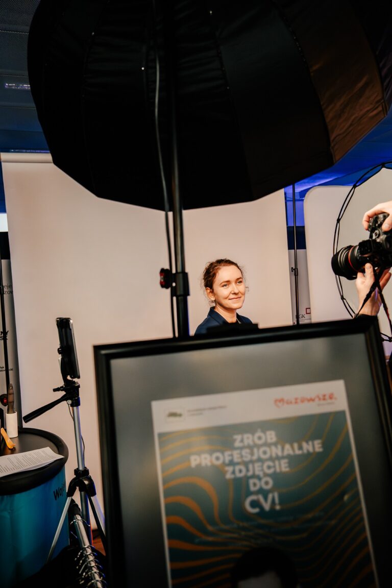 Na zdjęciu widać stanowisko fotograficzne, gdzie kobieta pozuje do zdjęcia portretowego. Na pierwszym planie znajduje się plakat z napisem „Zrób profesjonalne zdjęcie do CV!”. W tle widać oświetlenie studyjne oraz fotografa wykonującego zdjęcia.