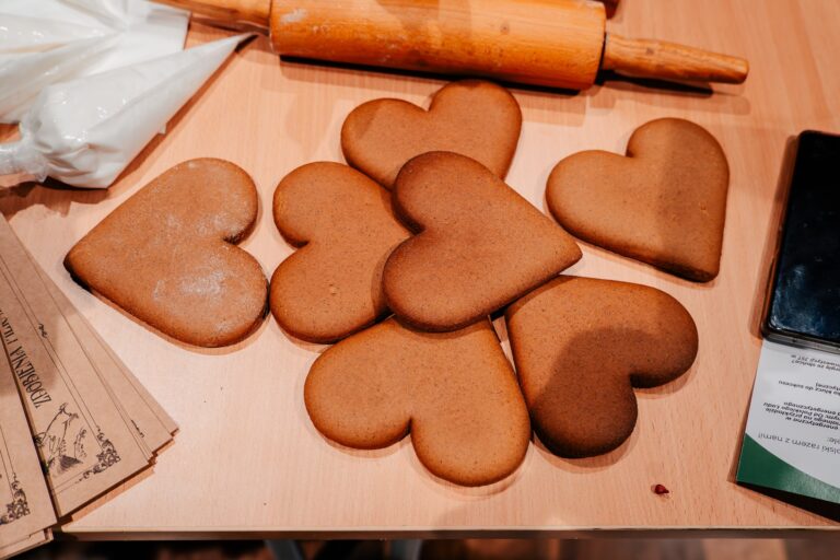 Na stole leży kilka pierników w kształcie serca, obok znajduje się wałek do ciasta i worki cukiernicze z lukrem. Scena przygotowana do dekorowania ciastek.