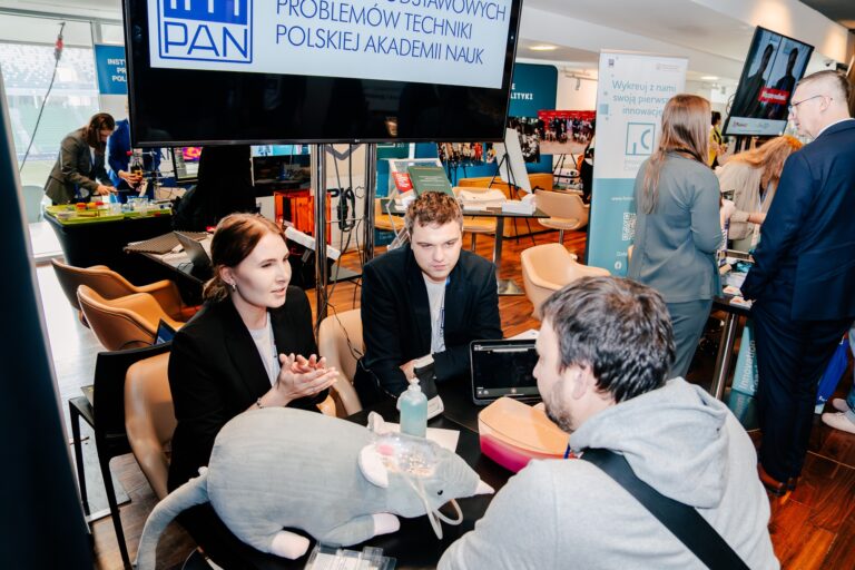 Stoisko Instytutu Podstawowych Problemów Techniki Polskiej Akademii Nauk podczas 15. Forum Rozwoju Mazowsza. Uczestnicy rozmawiają przy stole z prezentowanymi materiałami edukacyjnymi i sprzętem demonstracyjnym.