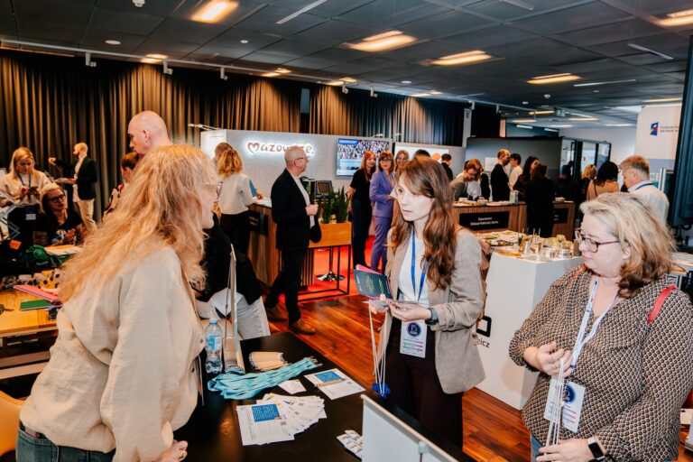 Uczestnicy 15. Forum Rozwoju Mazowsza rozmawiają przy stoisku programu Innovation Coach. W tle widoczne są inne ekspozycje, m.in. Mazowsza i instytucji wspierających rozwój regionu