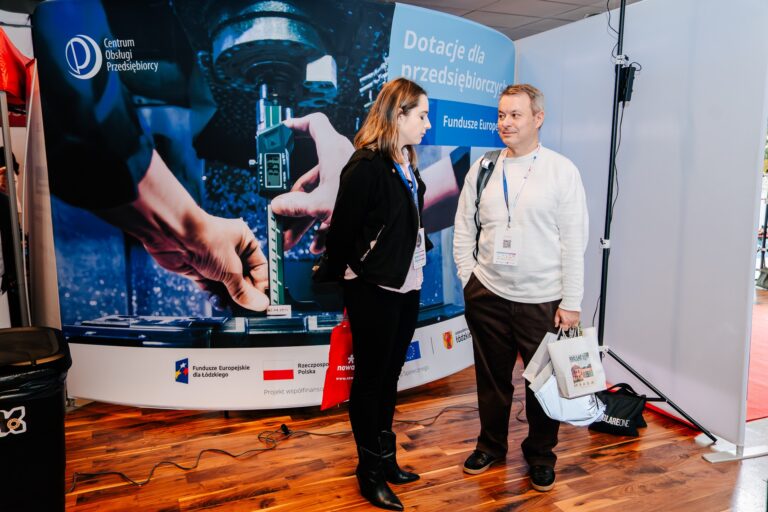 Dwie osoby rozmawiają przy stoisku Centrum Obsługi Przedsiębiorcy podczas 15. Forum Rozwoju Mazowsza. W tle widoczny jest baner promujący dotacje dla przedsiębiorców z Funduszy Europejskich.