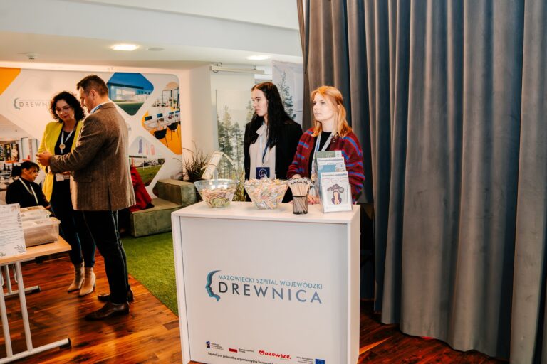 Stoisko Mazowieckiego Szpitala Wojewódzkiego Drewnica podczas 15. Forum Rozwoju Mazowsza. Dwie kobiety stoją przy ladzie informacyjnej z materiałami edukacyjnymi i gadżetami promocyjnymi.