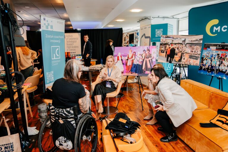 Uczestniczki 15. Forum Rozwoju Mazowsza rozmawiają przy stoisku programu Innovation Coach oraz Instytutu Podstawowych Problemów Techniki PAN. W tle widoczne są plansze ze zdjęciami projektów i wydarzeń kulturalnych.