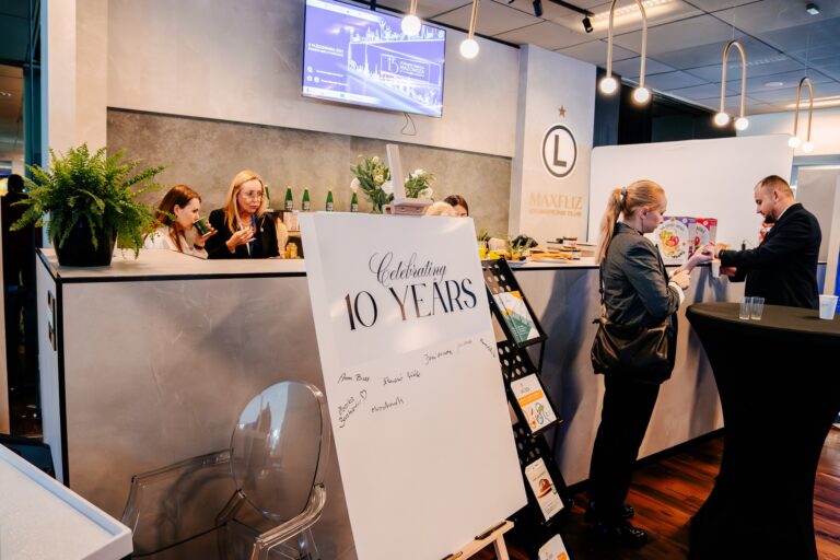 Stoisko Unimos Alliance podczas 15. Forum Rozwoju Mazowsza. Na pierwszym planie tablica z napisem „Celebrating 10 Years”, a w tle uczestnicy rozmawiają przy ladzie z napojami i materiałami promocyjnymi.