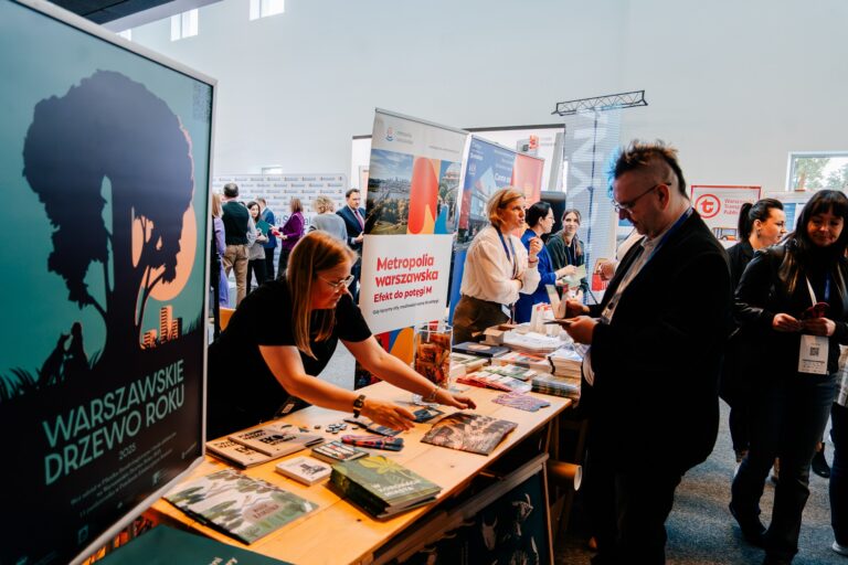 Stoisko Metropolii Warszawskiej podczas 15. Forum Rozwoju Mazowsza. Przedstawicielka prezentuje materiały informacyjne i gadżety promujące inicjatywy regionalne, obok widoczny plakat „Warszawskie Drzewo Roku 2025
