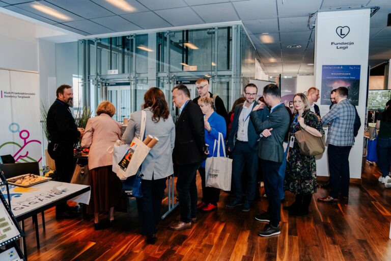 Uczestnicy 15. Forum Rozwoju Mazowsza rozmawiają i oglądają stoiska wystawców w przestrzeni z przeszklonymi windami. Część osób trzyma torby z materiałami informacyjnymi, a w tle widoczne są banery Klubu Przedsiębiorczości Dzielnicy Targówek.