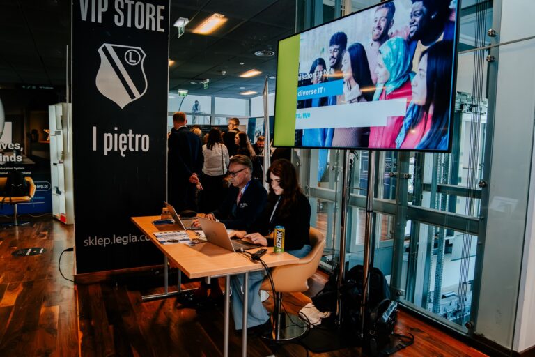 Dwie osoby siedzą przy stole z laptopami podczas 15. Forum Rozwoju Mazowsza. Obok nich duży ekran z prezentacją i grupa uczestników rozmawiających w tle.