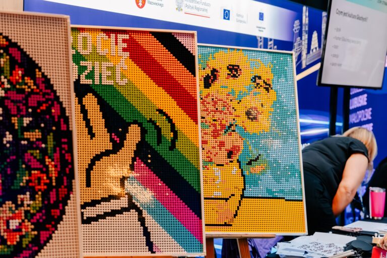 Kolorowe mozaiki z koralików przedstawiające różne wzory, w tym kwiaty i dłoń na tle tęczy, zaprezentowane na stoisku Domu Kultury Świt podczas 15. Forum Rozwoju Mazowsza.