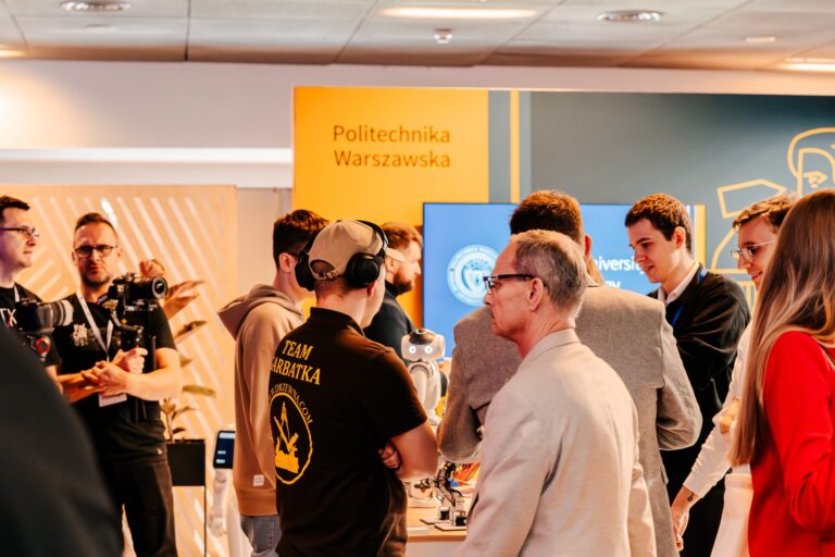 Stoisko Politechniki Warszawskiej podczas 15. Forum Rozwoju Mazowsza. Grupa uczestników ogląda prezentację z robotem humanoidalnym, a w tle widoczny jest ekran z logotypem uczelni.