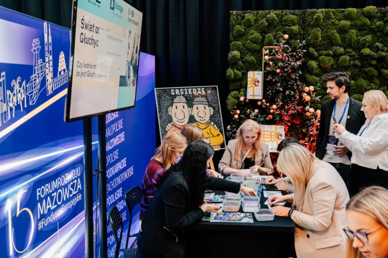 Stoisko Domu Kultury Świt podczas 15. Forum Rozwoju Mazowsza. Uczestnicy wykonują kolorowe mozaiki z koralików przy stole warsztatowym. W tle ekran z napisem „Świat oczami Głuchych” oraz obraz przedstawiający postacie z serialu „Sąsiedzi”.