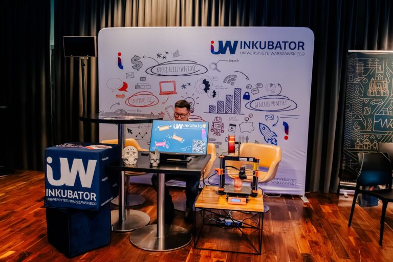 Stoisko Inkubatora Uniwersytetu Warszawskiego podczas 15. Forum Rozwoju Mazowsza. Przy stole siedzi osoba z laptopem, obok znajduje się drukarka 3D i elementy promujące innowacje oraz przedsiębiorczość.
