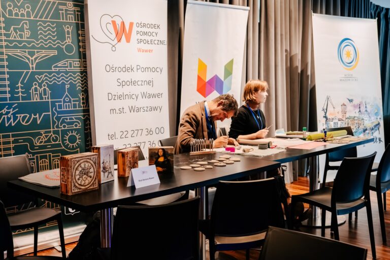 Stoisko Ośrodka Pomocy Społecznej Dzielnicy Wawer podczas 15. Forum Rozwoju Mazowsza. Dwie osoby przygotowują materiały i prezentują rękodzieło artystyczne na stole z oznaczeniem „Klub Seniora Wawer