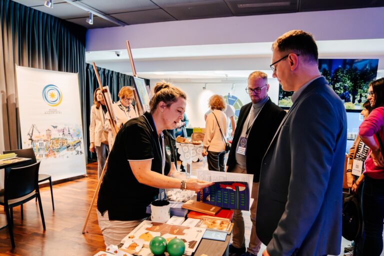 Stoisko dzielnicy Wawer podczas 15. Forum Rozwoju Mazowsza. Kobieta prezentuje materiały edukacyjne i informacyjne odwiedzającym, w tle widać inne stoiska oraz uczestników wydarzenia.