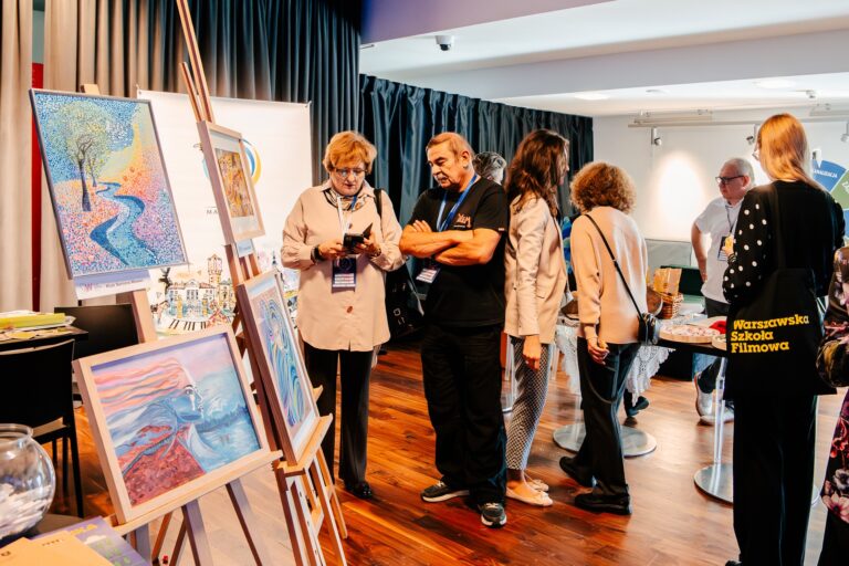 Seniorzy z dzielnicy Wawer prezentują swoje prace plastyczne podczas 15. Forum Rozwoju Mazowsza. Na sztalugach stoją kolorowe obrazy, a uczestnicy wydarzenia oglądają wystawę i rozmawiają
